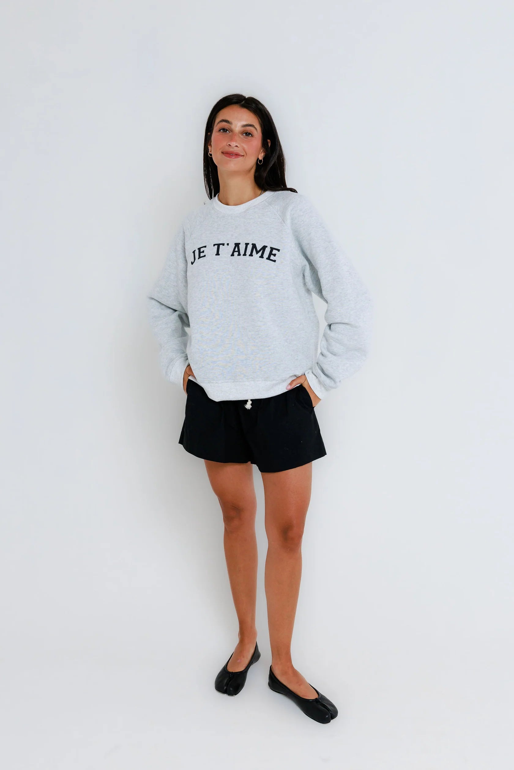 Woman wearing a lightweight white raglan crewneck sweatshirt, black shorts, and black flats on a plain white background.