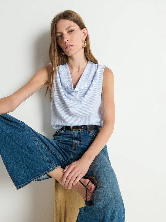 Woman in a striped cowl-neck tank top and wide-leg denim jeans sits on a stool against a plain white background.