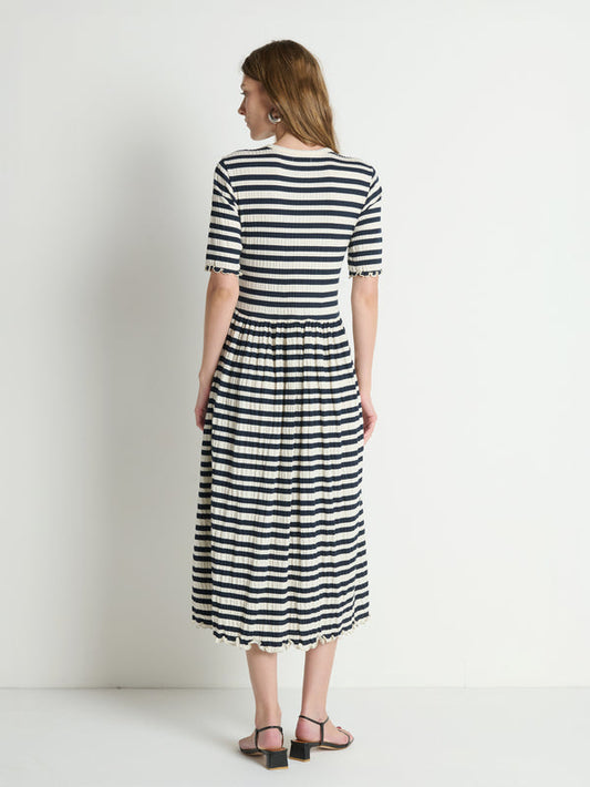 Woman wearing striped crinkle midi dress stands barefoot, facing a white wall.