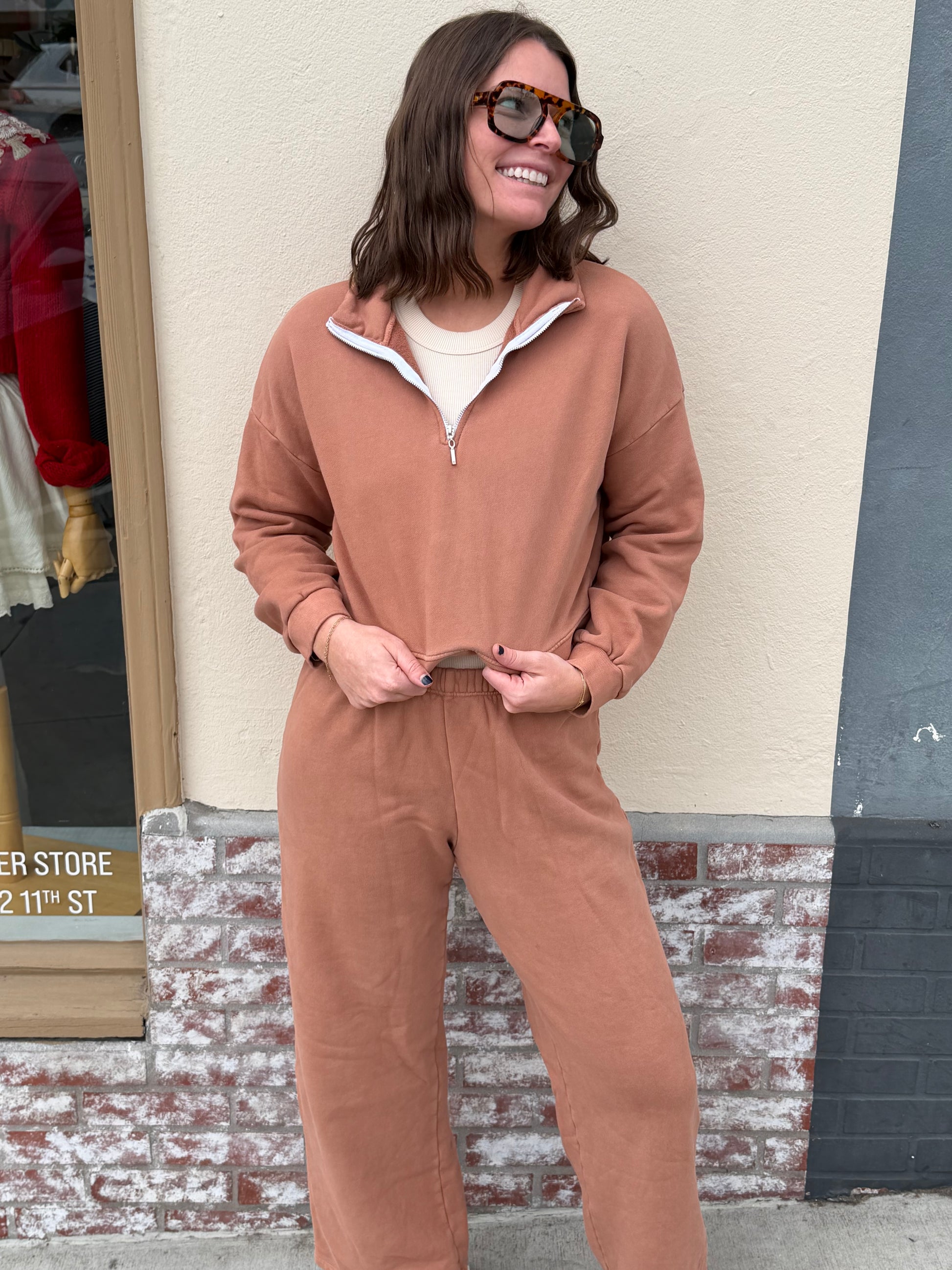 Woman wears a birch-colored zip-up jacket with sunglasses, standing outside a store.