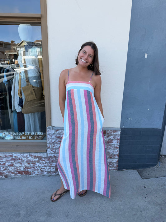 Woman wearing a striped multicolor dress stands next to a store window and against a white-gray wall.