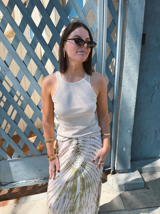 Woman in sunglasses wears a beige tank top and patterned skirt, standing by a blue lattice fence.