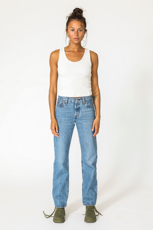 Woman wearing white tank top and blue jeans with green shoes, standing front-facing against a plain white background.
