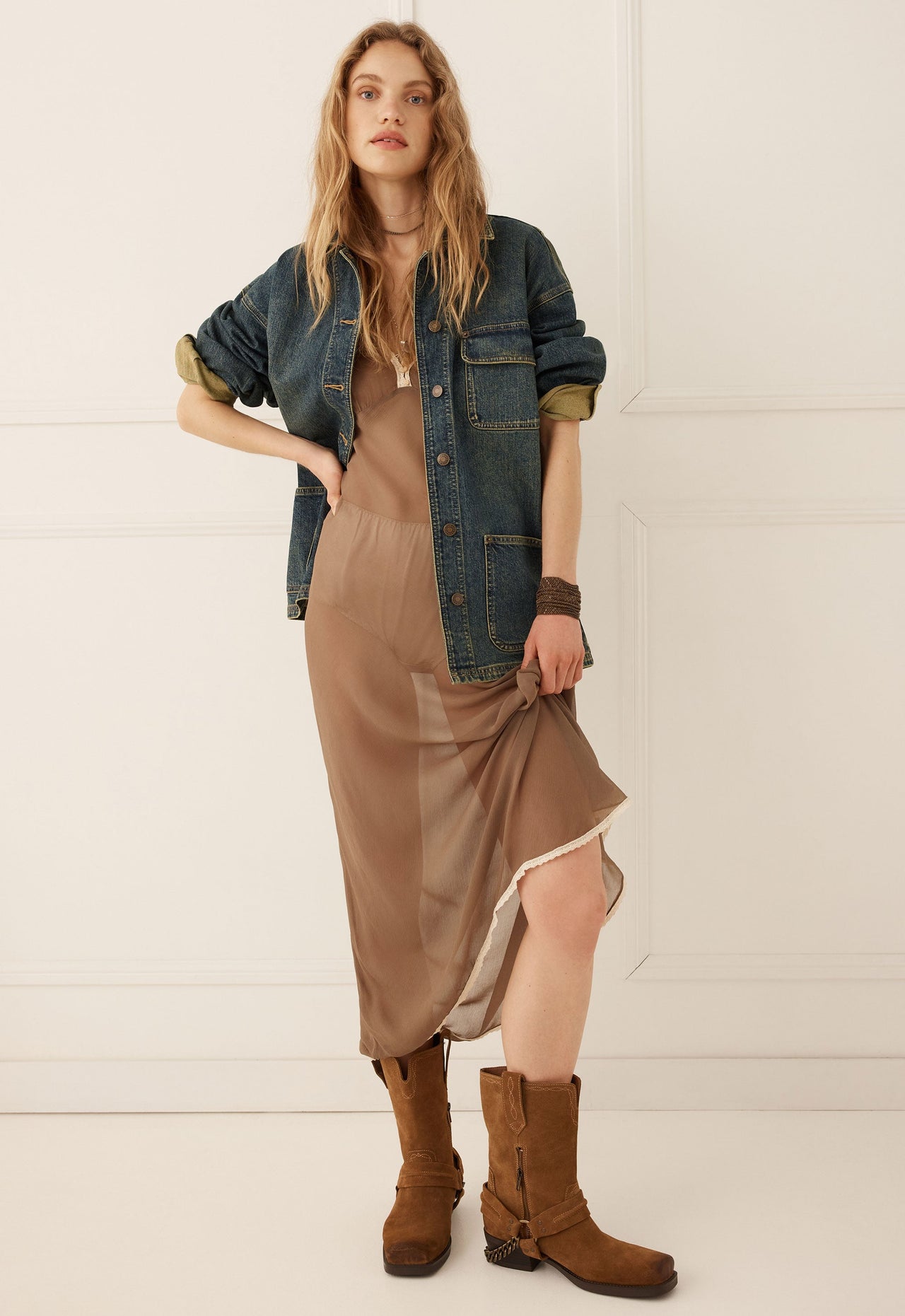 Woman wearing a sheer brown dress, denim jacket in indigo, and boots stands in a minimalist room, lifting her dress.
