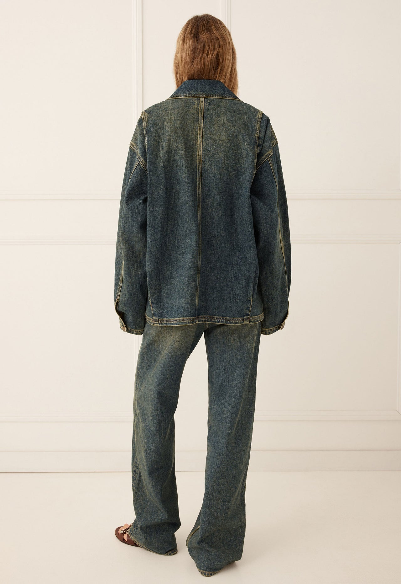 Person faces away in a minimal room, wearing a denim jacket in dirty indigo and matching jeans.