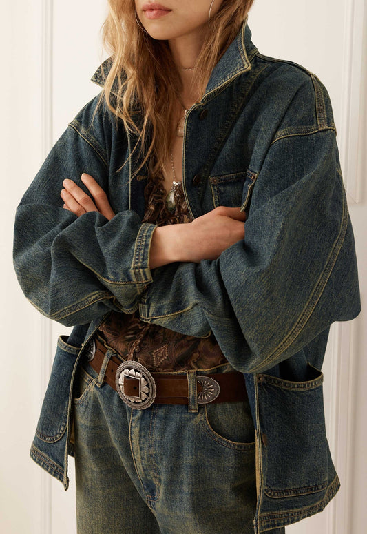 Woman in indigo denim jacket and jeans with arms crossed, wearing a brown belt with a silver buckle.