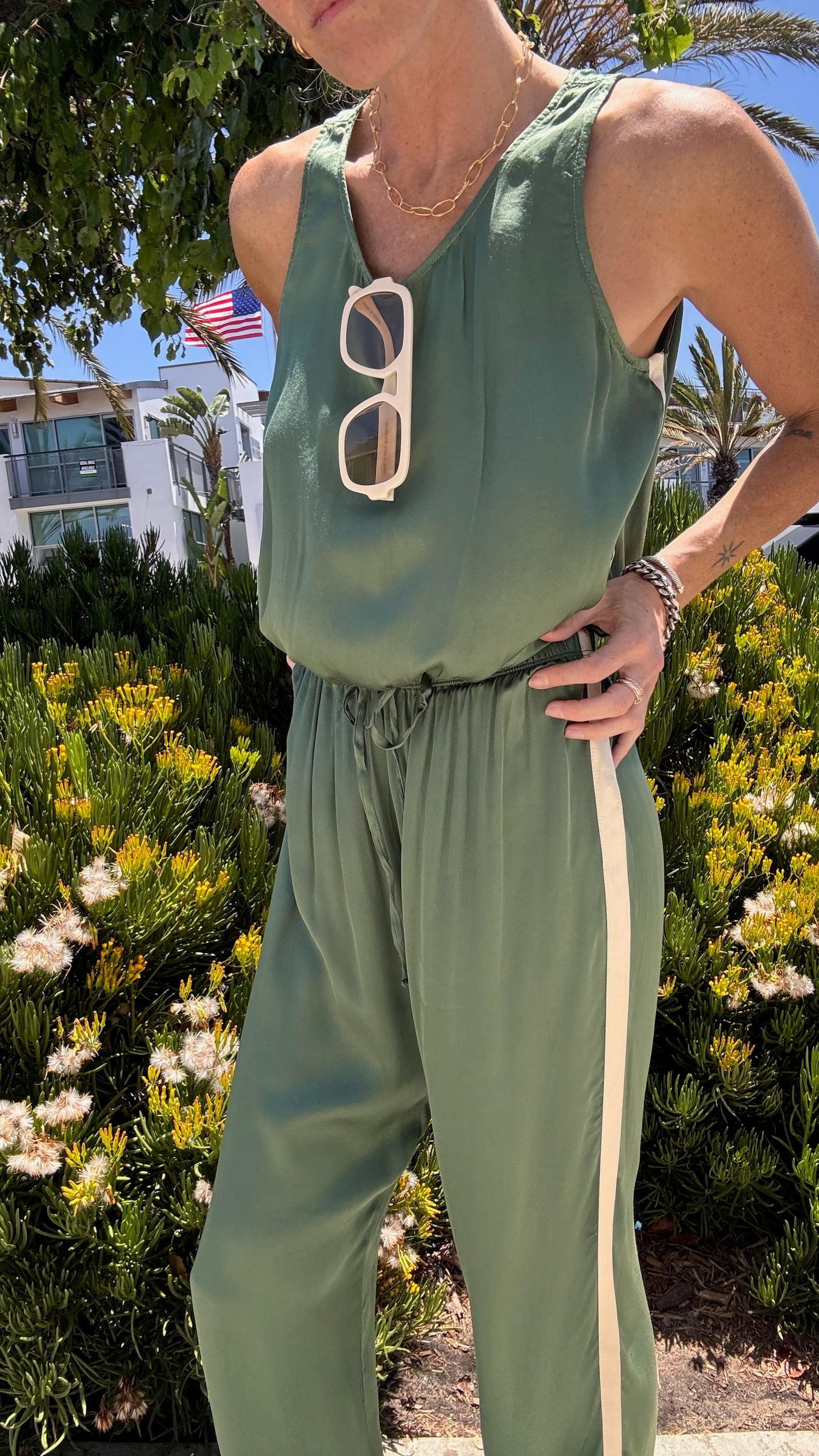 Person outdoors in celadon jade track pants and white sunglasses, surrounded by dense greenery.