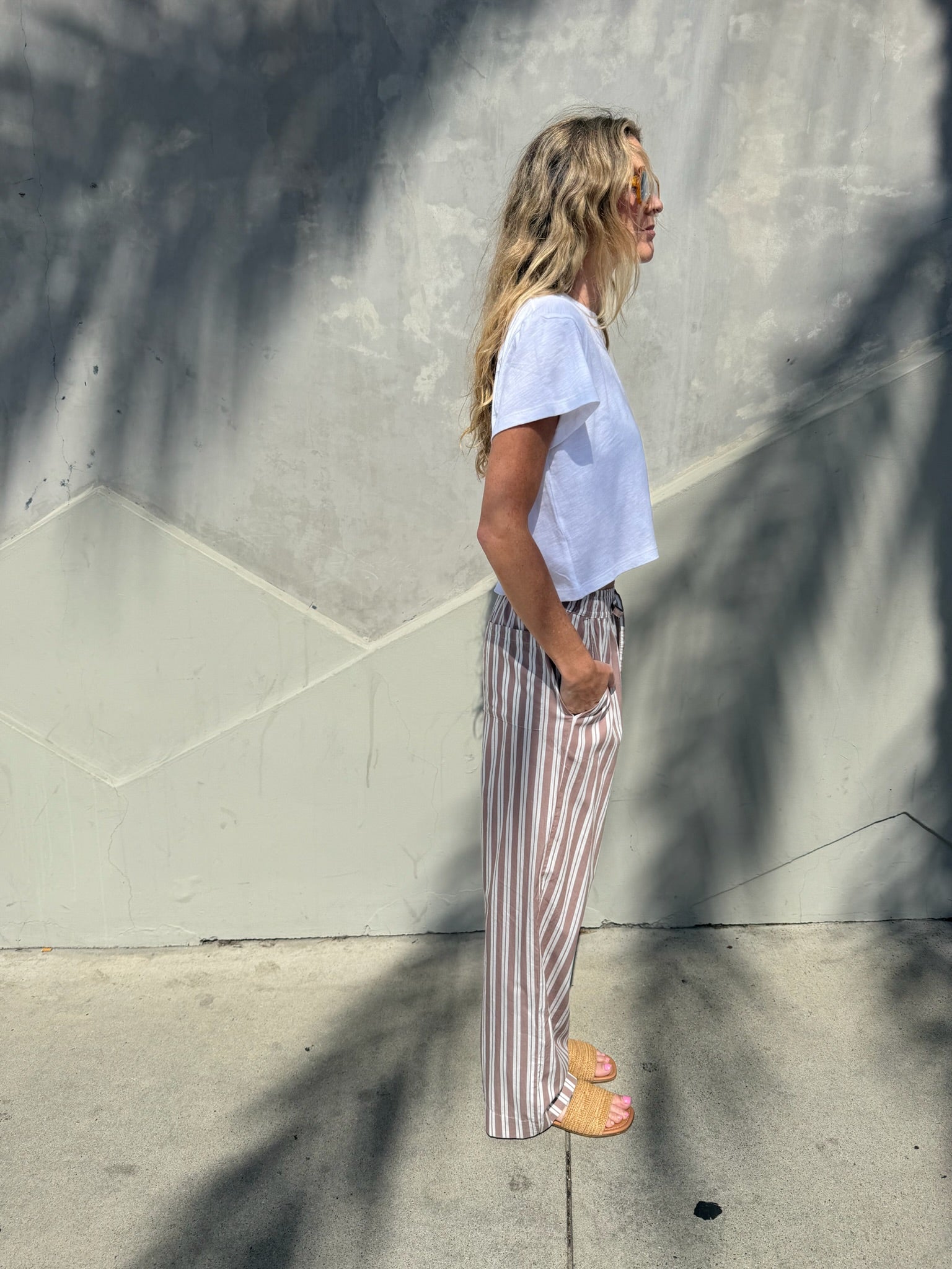 Woman wears wide-leg lounge pants in stucco color, standing on a sunny sidewalk under palm frond shade.