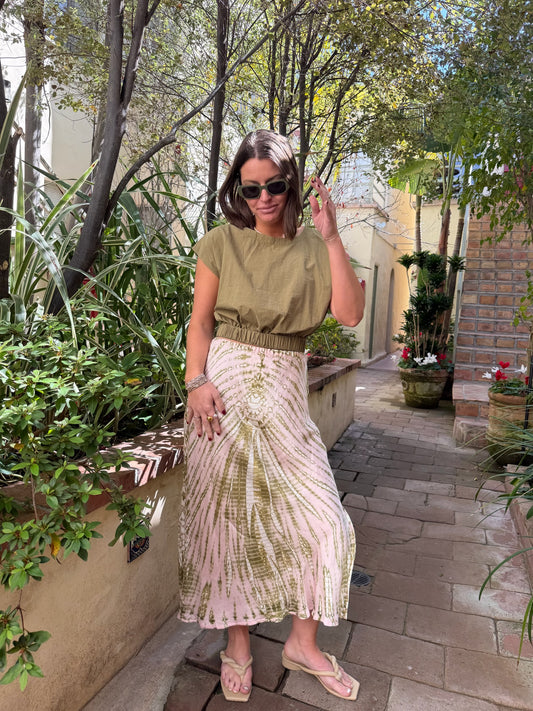 Woman with sunglasses wearing a green top and tie-dye skirt stands on a garden path.