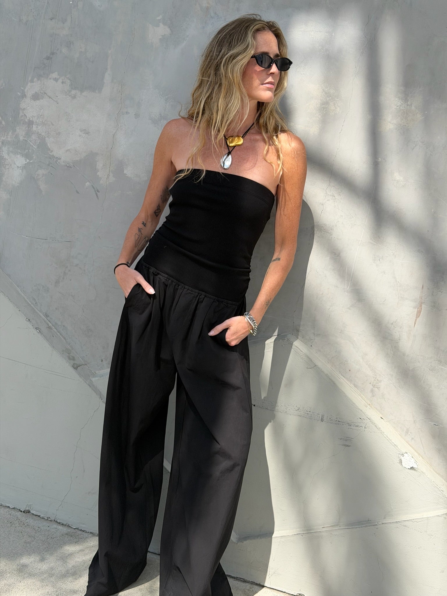 Woman in black jumpsuit with chunky necklace and sunglasses stands by a gray wall.