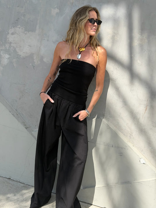 Woman in black jumpsuit with chunky necklace and sunglasses stands by a gray wall.