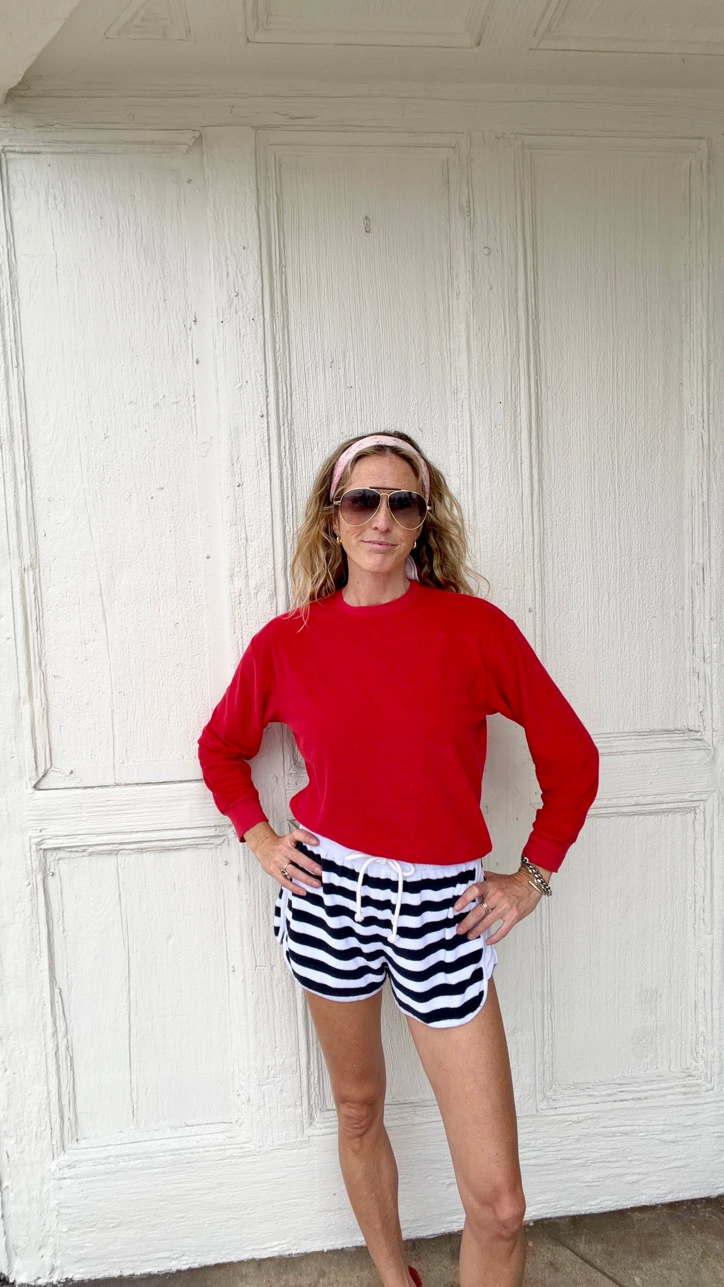 Woman in red sweater and white/navy athletic shorts stands with hands on hips in front of a white wooden door.