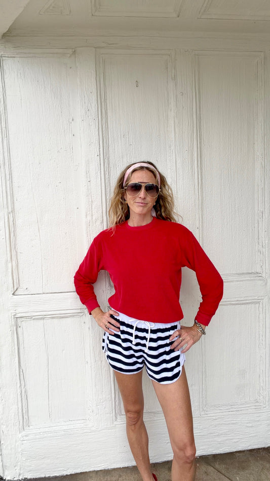 Woman in red sweater and white/navy athletic shorts stands with hands on hips in front of a white wooden door.
