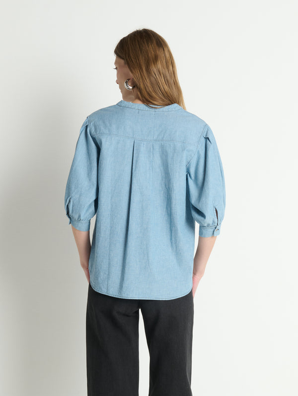 Back view of a woman in a chambray blue Emmett top and black pants, standing against a plain white background.