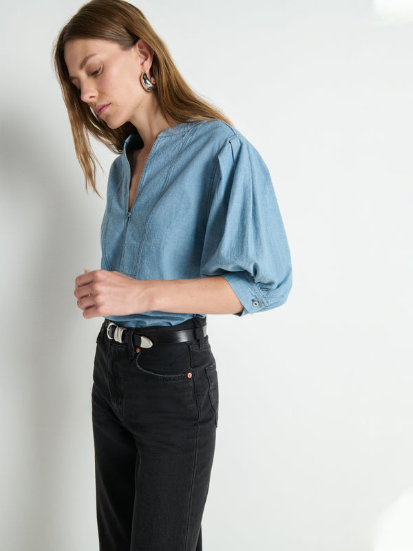 Woman in a chambray button-up top and black jeans stands against a white background, looking down.