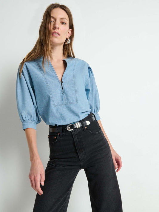 Woman in chambray Emmett top and high-waisted black jeans, posed in front of a white wall.