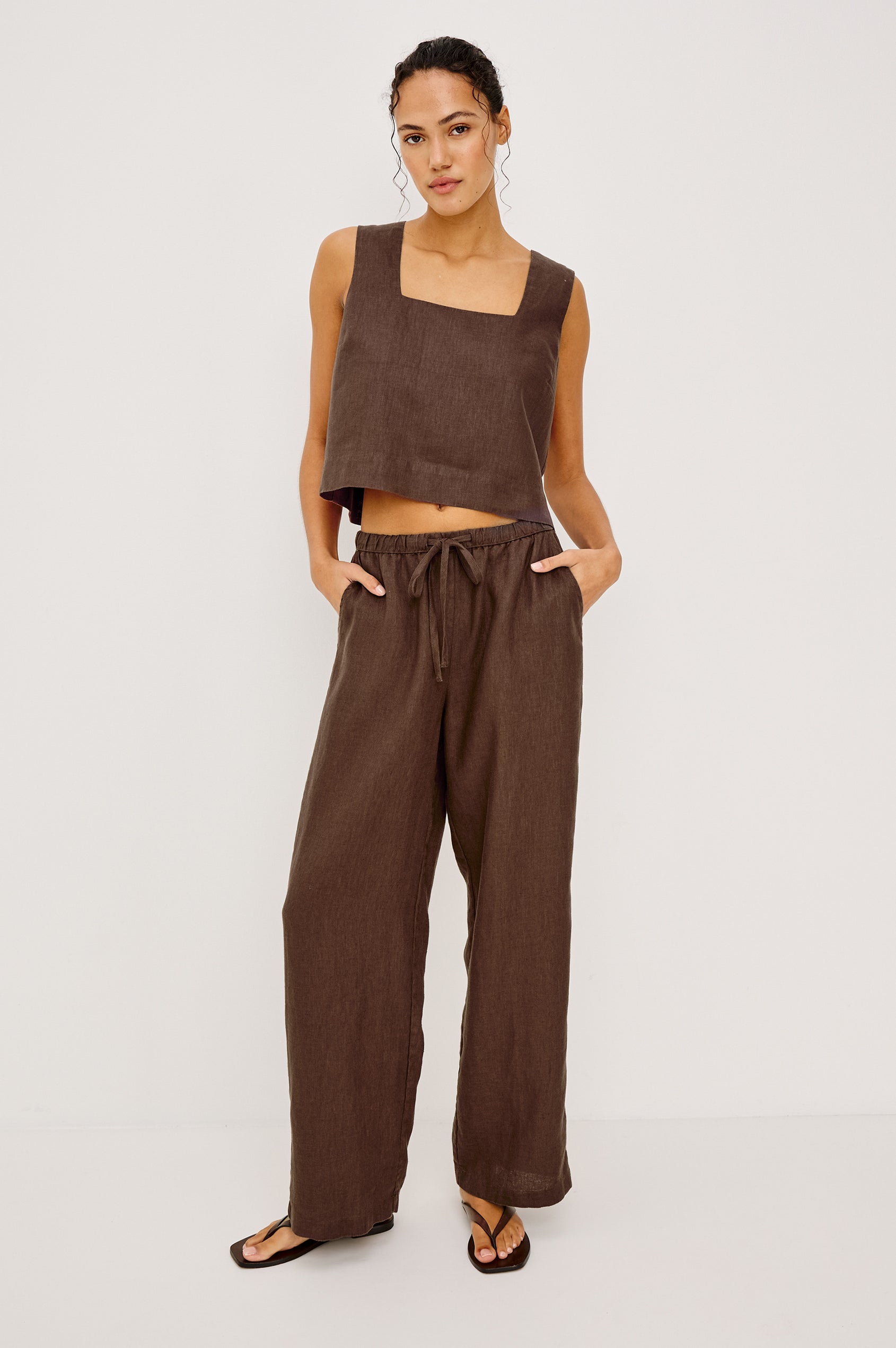 Woman in espresso-colored pants and a brown sleeveless crop top stands against a plain white background.
