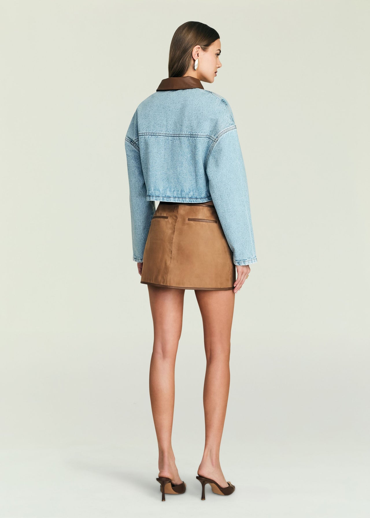 Woman facing away in cropped denim jacket, tan suede mini skirt, and brown heels. Casual outfit suitable for outings.