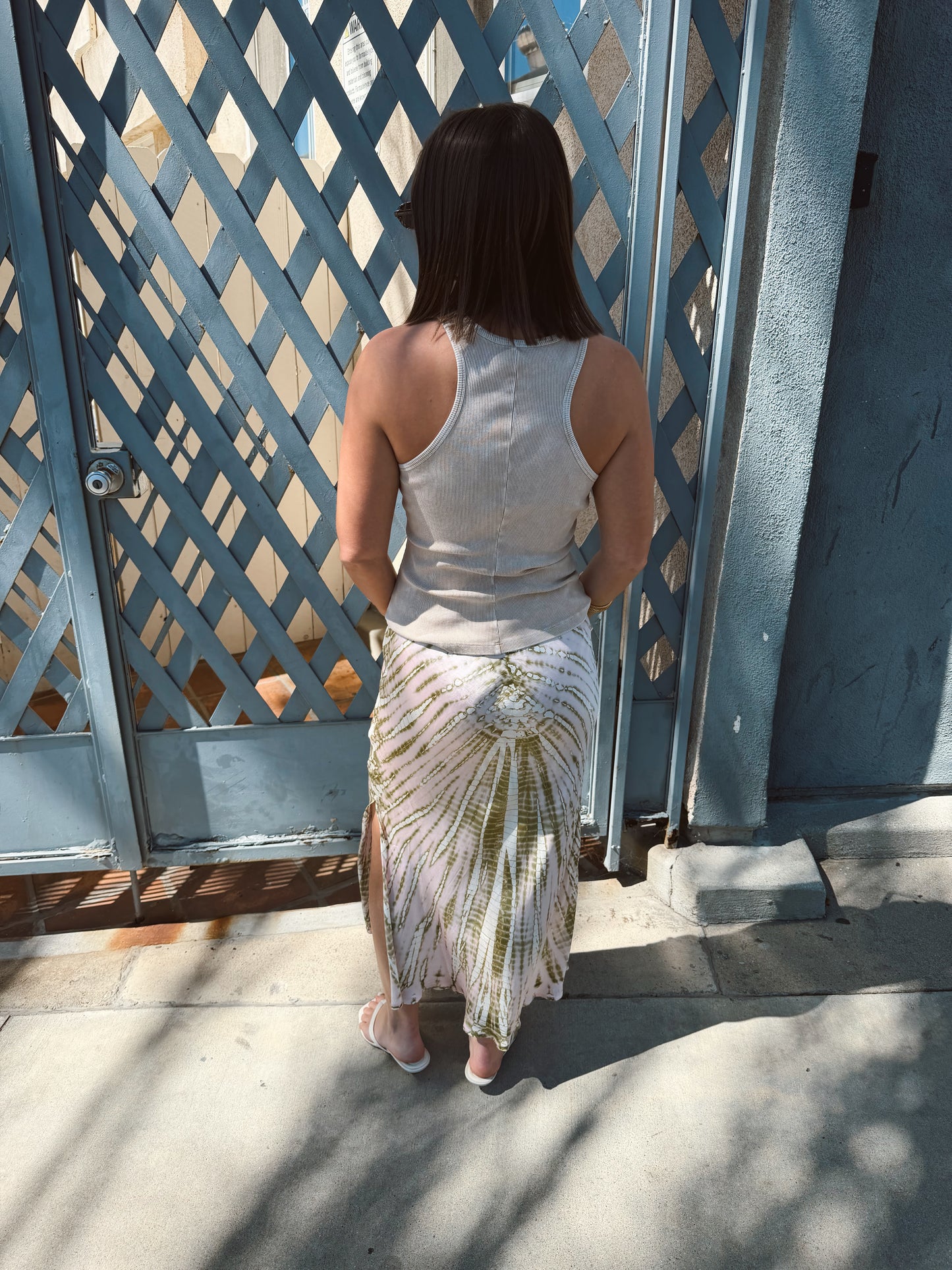 Woman in a sandstone-colored tank top stands on sidewalk, facing a blue lattice gate.