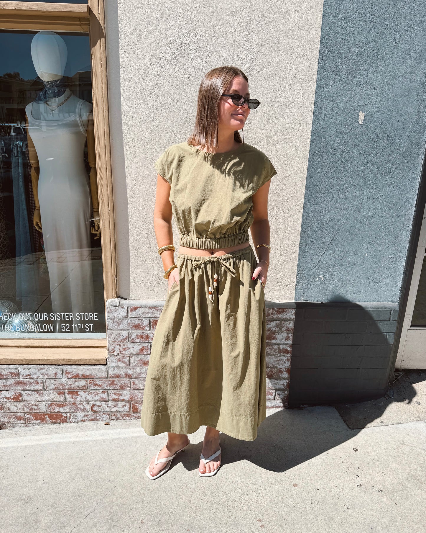 Woman in sunglasses wears a dark olive long-sleeve top, suitable for casual outdoor wear, standing on a sunny sidewalk.