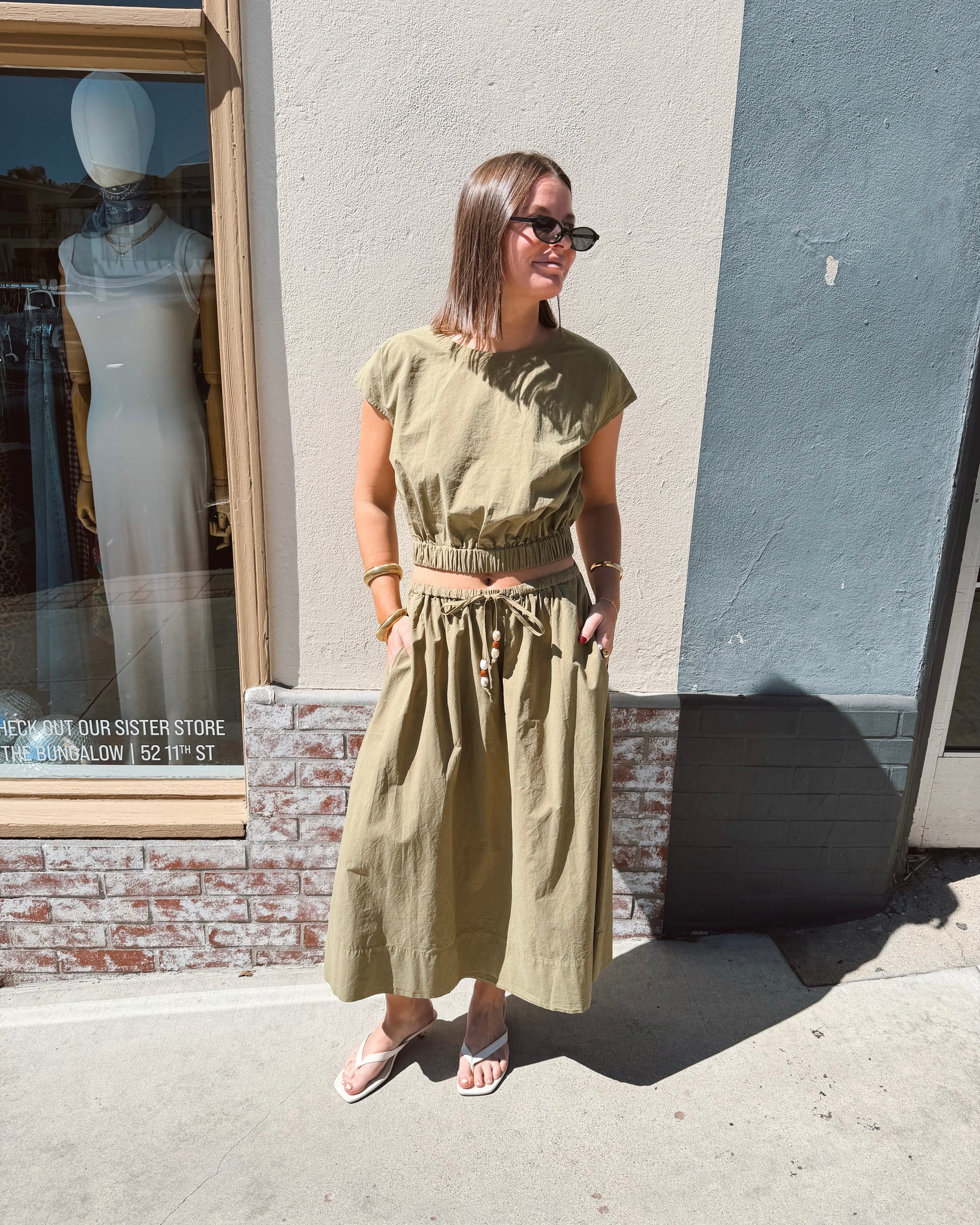 Woman in sunglasses wears a dark olive long-sleeve top, suitable for casual outdoor wear, standing on a sunny sidewalk.