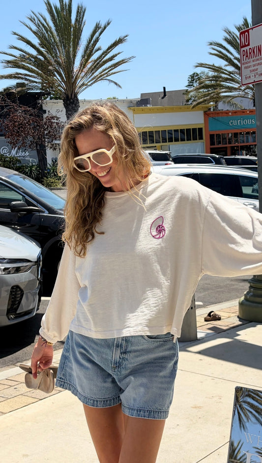 Woman in pearl-colored Seashell Sculpture sweatshirt, sunglasses, and denim shorts stands on a sunny sidewalk.