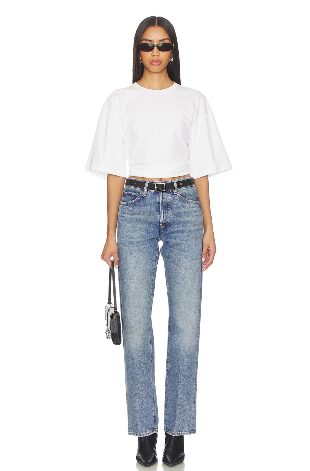 Woman wears blue jeans, a white oversized t-shirt, black boots, sunglasses, and carries a small black purse.
