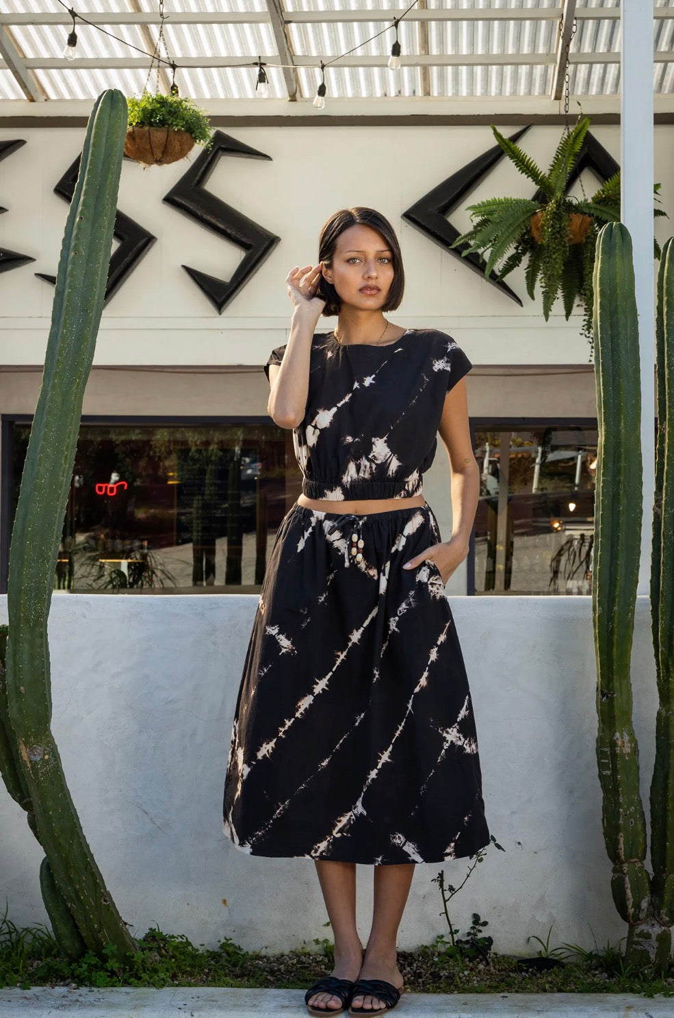 Woman in a tie-dye top stands between cacti outside a modern building. Cotton blend, casual wear.