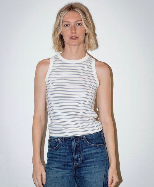 Woman in blue striped linen tank top and jeans stands against a white wall.