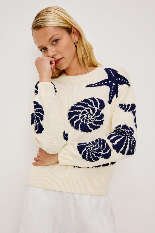 Woman wearing a cream knit sweater with seashell print and a white skirt, hand resting near her face.