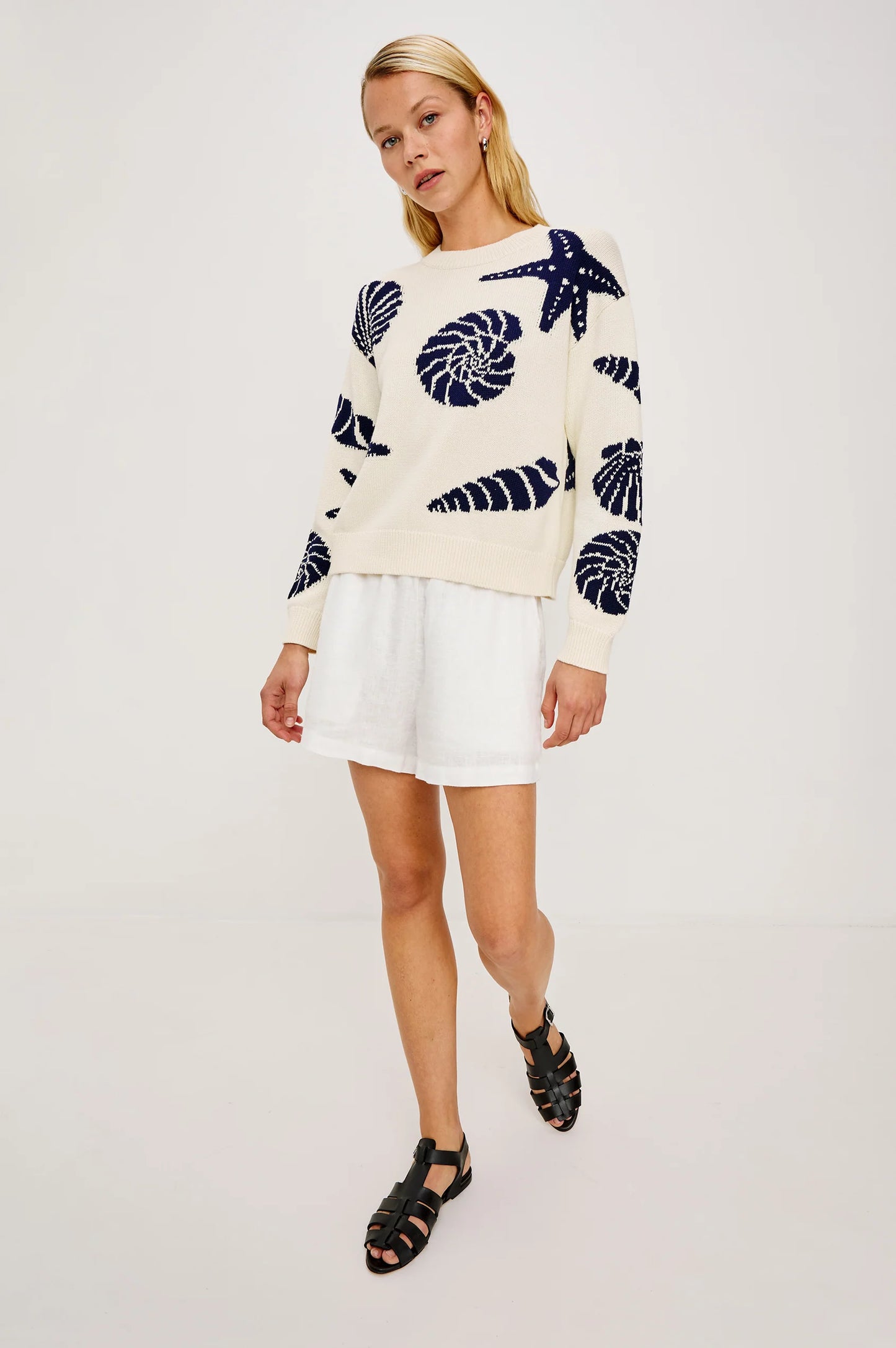 Woman in a light knit seashell-pattern sweater with white shorts and black sandals, standing against a plain background.
