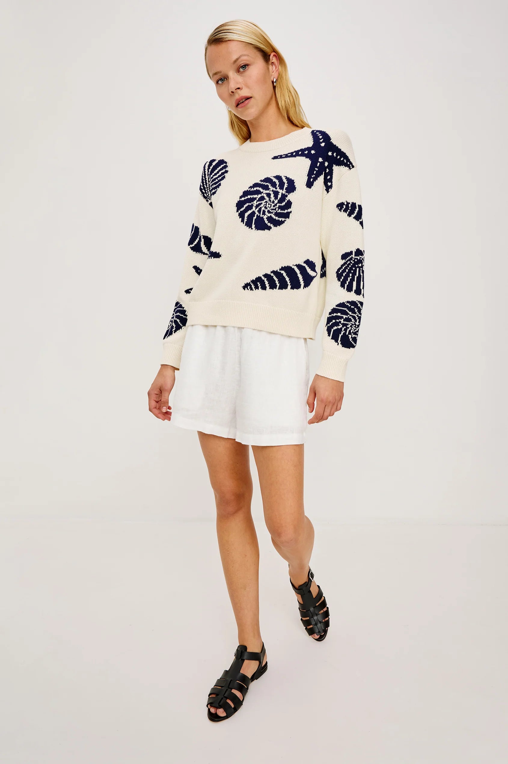 Woman in a light knit seashell-pattern sweater with white shorts and black sandals, standing against a plain background.