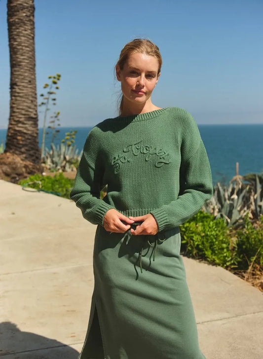 Woman wears a green crop boat neck sweater by palm trees near the ocean, showing casual knitwear for coastal settings.