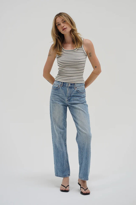 Woman wears a striped grey tank top, blue jeans, and sandals standing against a plain white background.