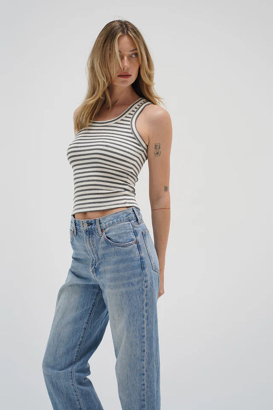 Woman in a striped grayscale tank top and blue jeans stands against a light background, looking to the side.