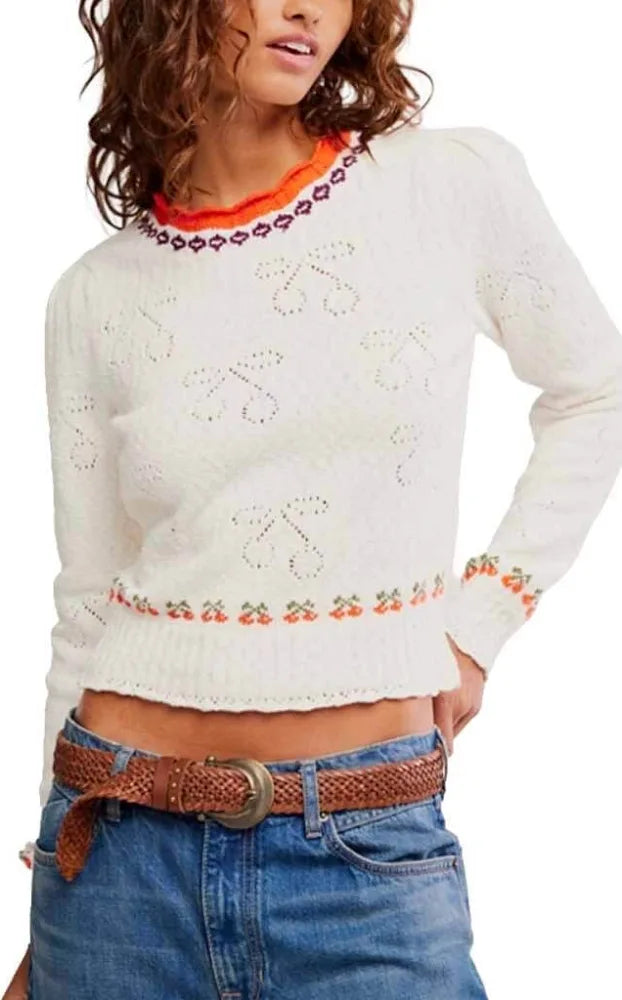Woman in a cream knit pullover, blue jeans, and brown braided belt stands with hand on hip.
