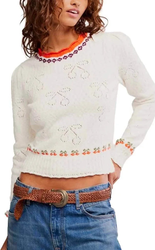 Woman in a cream knit pullover, blue jeans, and brown braided belt stands with hand on hip.