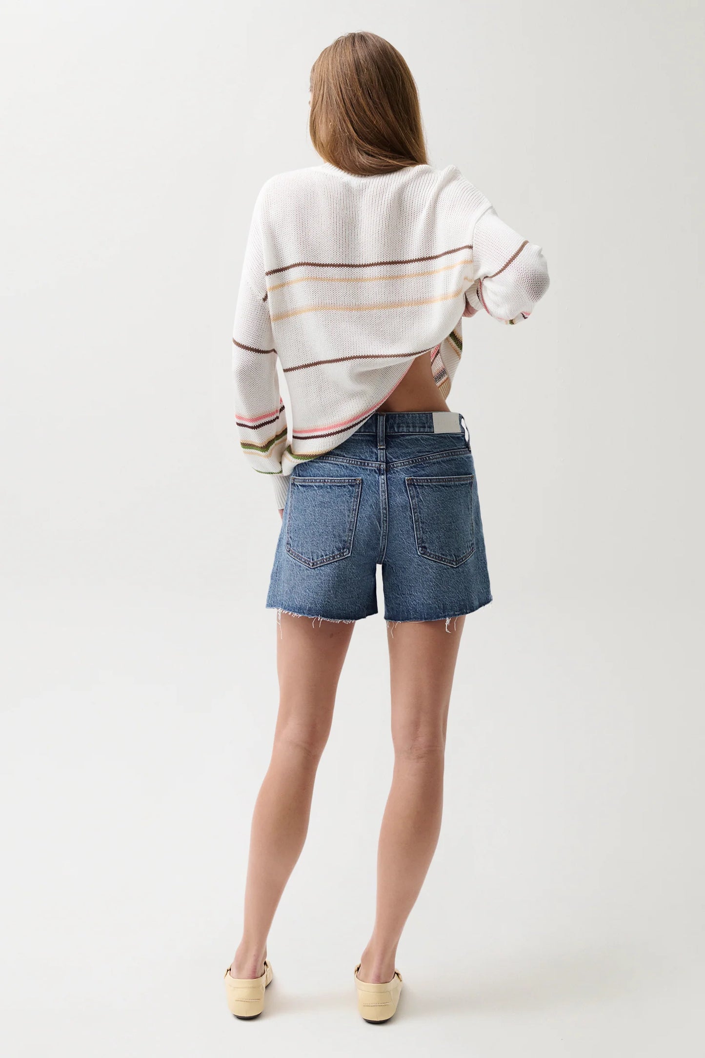Woman shown from behind in a white sweater, light wash denim cutoff shorts, and beige slip-on shoes.