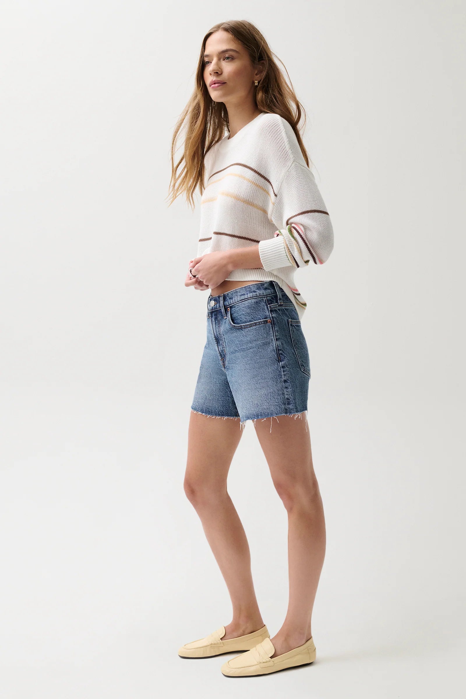 Woman in profile wearing a white sweater, light denim cut-off shorts, and beige loafers.