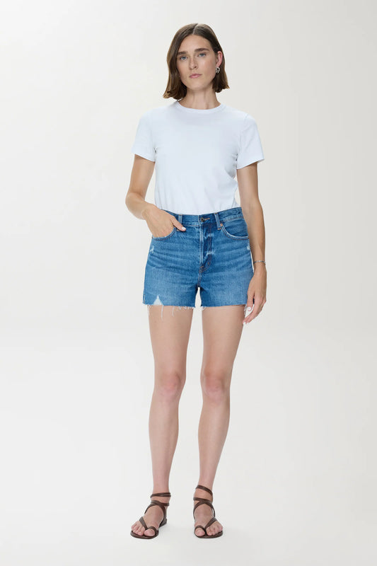 Woman wearing light-wash high rise denim cut-off shorts and a white cotton t-shirt, standing before a plain background.