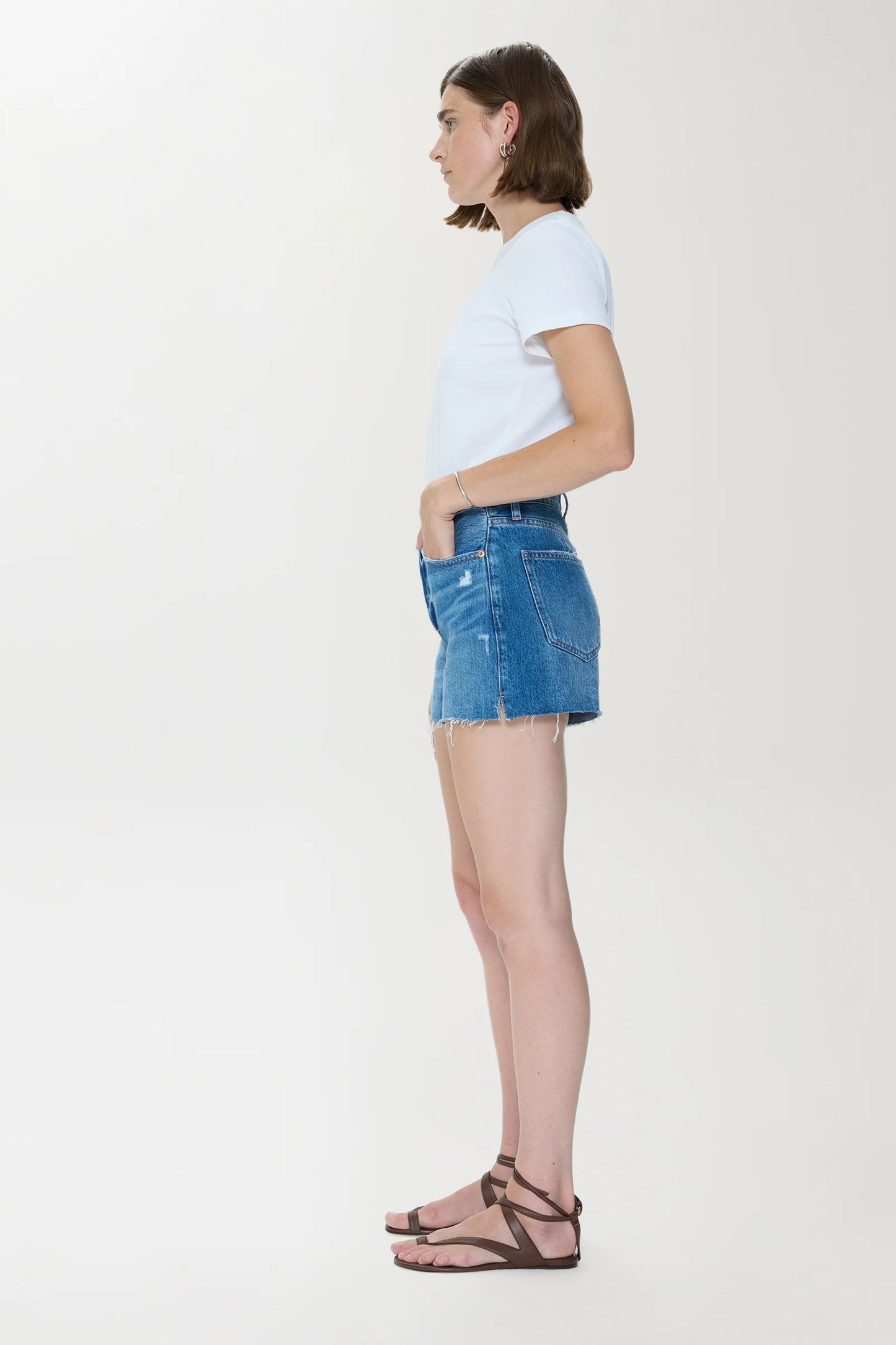 Woman in white t-shirt, light denim high-rise cut off shorts, and brown sandals stands in profile against plain background.
