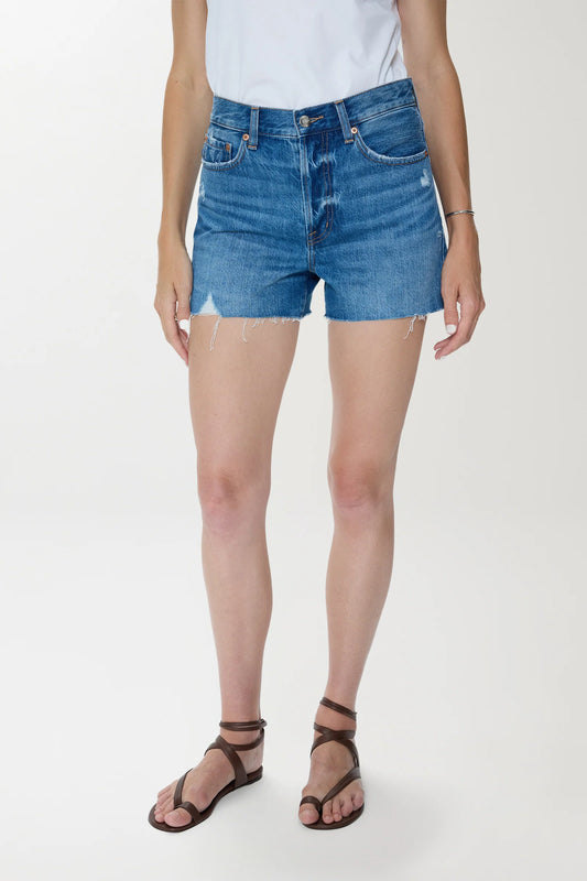 Woman in light denim high-rise cutoff shorts and a white cotton t-shirt, standing against a plain background.