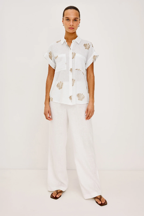 Woman in taupe seashell-patterned button-up shirt, white wide-leg pants, and brown sandals stands before a plain background.