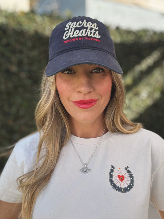 Woman with long blonde hair wears a white hat, heart-print shirt, and a silver eye-shaped necklace.