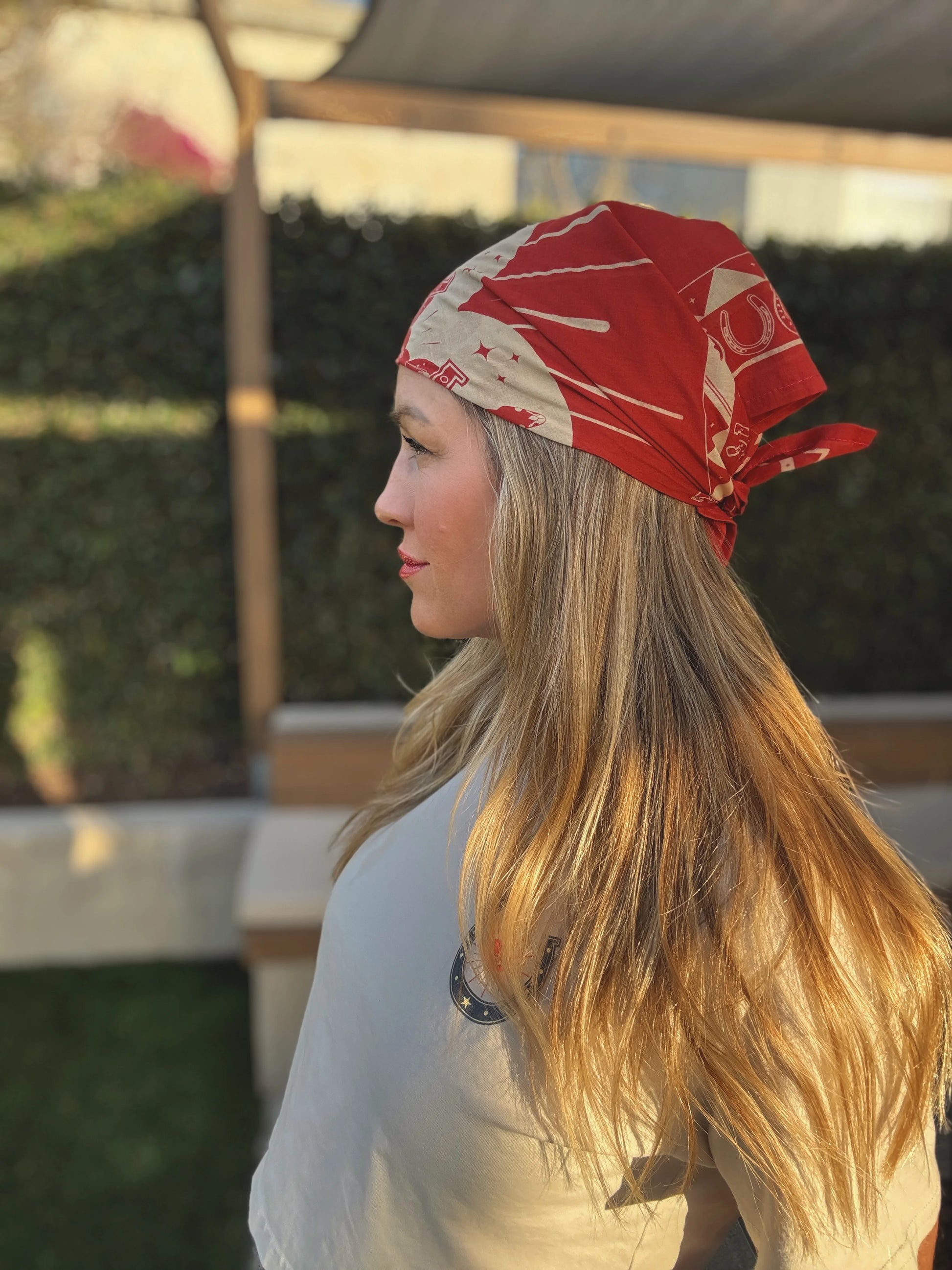 Woman with long blonde hair wears a patterned fabric bandana outdoors in sunlight, shown from the side.