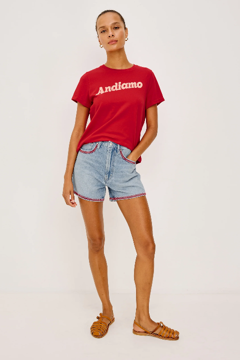 Woman wearing a red cotton crewneck T-shirt, denim shorts, and sandals against a plain white background.