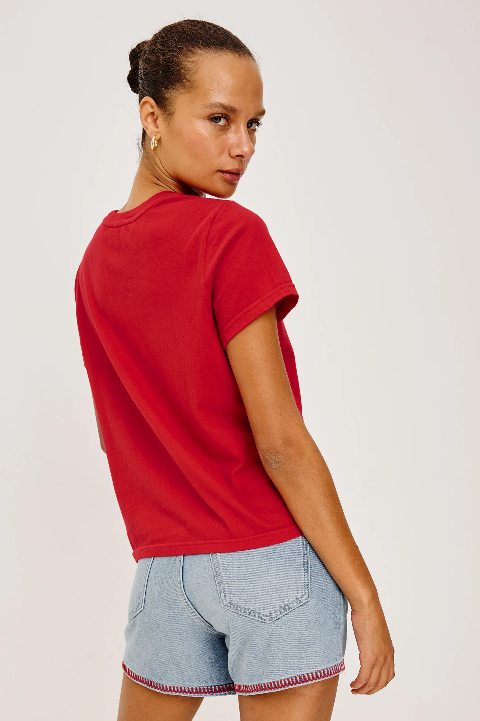 Woman in a red cotton crew neck tee and denim shorts, turned slightly, glancing over her shoulder.