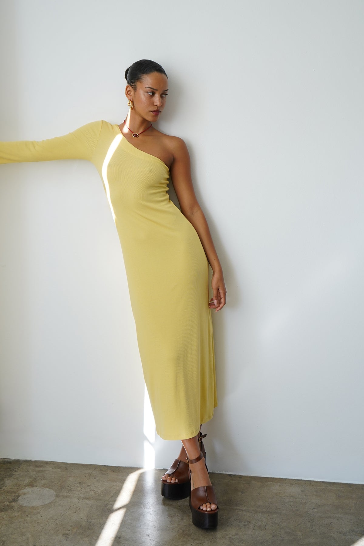 Woman in a raffia-colored ribbed one-shoulder dress stands by a white wall on a concrete floor.