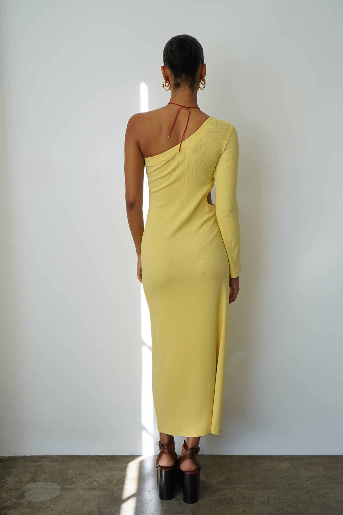 Back view of a woman wearing a raffia-colored ribbed one-shoulder dress, standing against a white wall.