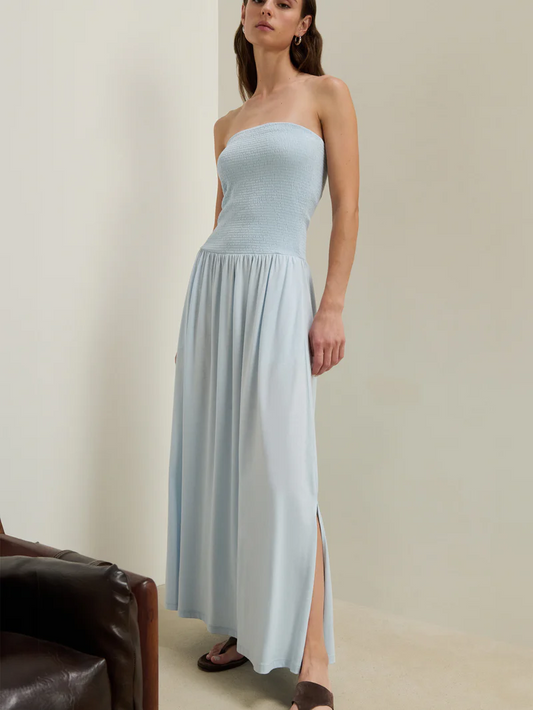 Woman wearing a light blue tube dress, standing indoors next to a wooden chair. Cotton fabric, casual or semi-formal use.